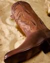 A close-up of a brown leather cowboy boot with horse-shaped stitching, resting on sandy ground.