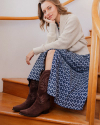 A woman wearing a light sweater, blue patterned skirt, and brown boots sits on wooden stairs, resting her head on her hand.