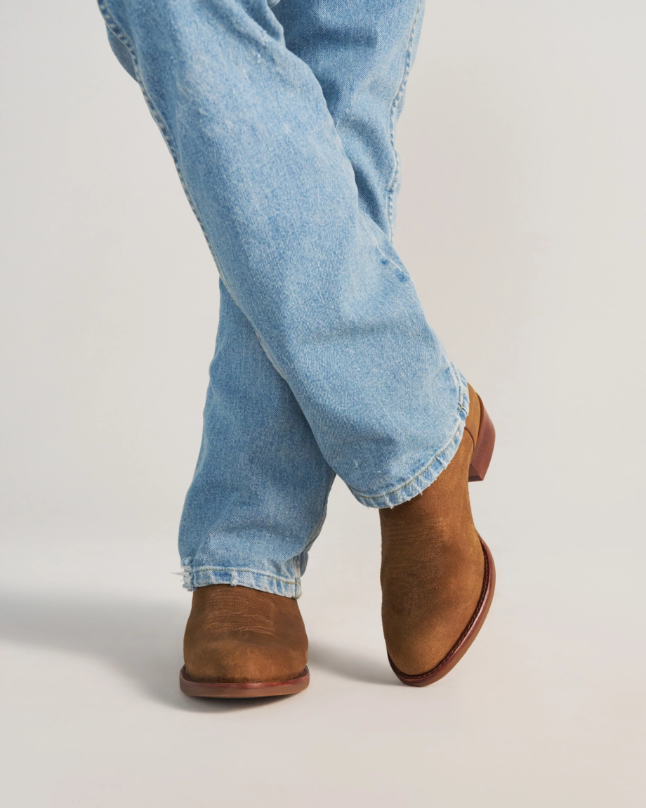 The Buck in Copper Cowhide. Person wearing light blue jeans and brown suede boots, standing with legs crossed on a plain white background, showcases subtle Copper Cowhide accents for a touch of rustic charm.