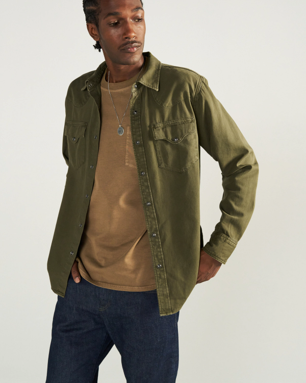A man with a thin mustache and braided hair wears an olive green button-down shirt over a tan pocket t-shirt, dark blue jeans, and a silver pendant necklace against a plain light background.