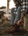 A person in jeans and a plaid shirt sits on a log, pulling on brown cowboy boots. Tattoos are visible on their arms and there are rocks in the background.