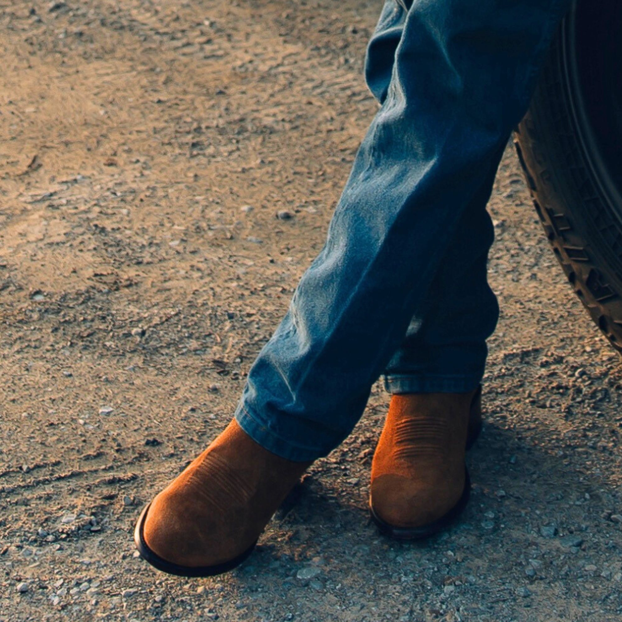 Person wearing blue jeans and brown boots stands with legs crossed on a gravel surface next to a vehicle tire.