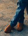Person wearing blue jeans and brown boots stands with legs crossed on a gravel surface next to a vehicle tire.