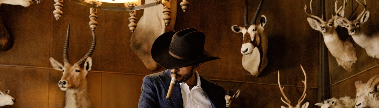 A man in a cowboy hat and suit stands by a pool table in a room decorated with wall-mounted animal trophies.