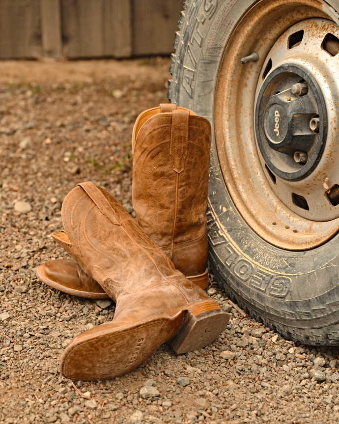 A pair of worn brown cowboy boots leaning against a dusty Jeep tire on a gravel surface.