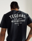 A person with braided hair wears a black T-shirt with white text on the back reading "Tecovas maker of fine Western Goods, Austin, Tex.