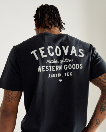 A person with braided hair wears a black T-shirt with white text on the back reading "Tecovas maker of fine Western Goods, Austin, Tex.