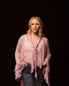 Woman in a pink blouse and jeans stands against a dark background, hands in pockets, with a calm expression.