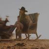 A woman in a cowboy hat and boots sits on a rocking chair outdoors with a can on the ground beside her; a blurred vehicle passes in the background.