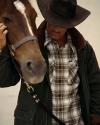 A person wearing a plaid shirt, jacket, and wide-brim hat stands close to a brown horse, gently touching its face.