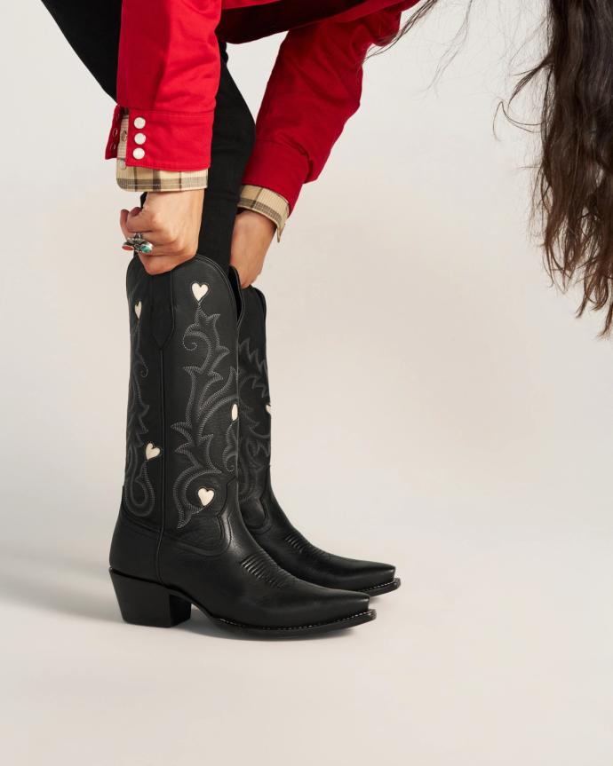 A person in a red shirt bends down to pull on black cowboy boots decorated with white heart-shaped accents.