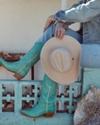 A person in jeans and teal cowboy boots sits on a concrete ledge, holding a beige hat over their knee, with rustic decor in the background.
