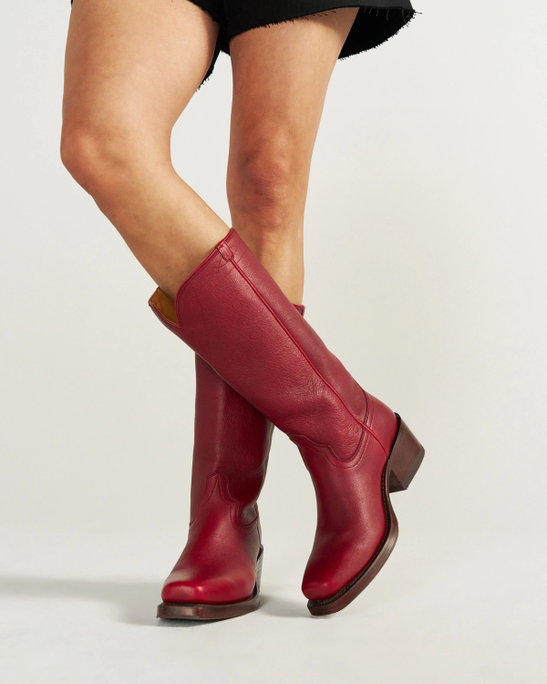 The Charlie in Scarlet Cowhide. Person wearing knee-high Scarlet Cowhide red leather boots with a low heel and a black skirt, standing against a plain white background.