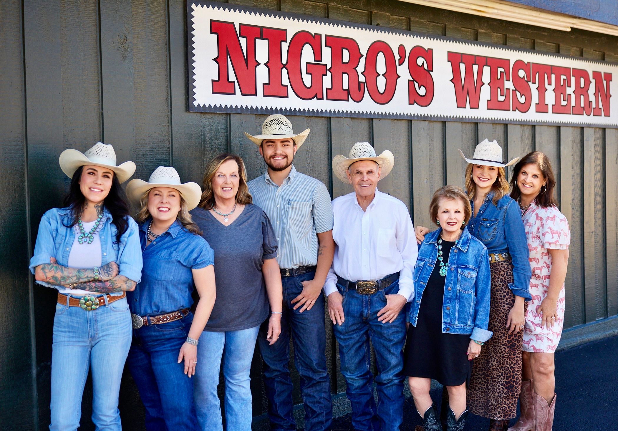 A group of eight people in western attire stand smiling in front of a building with a "NIGRO'S WESTERN" sign.