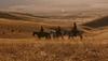 Three people ride horses across a dry, grassy field with hills and distant mountains in the background under a hazy sky.