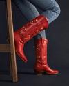 Person wearing blue jeans and red embroidered cowboy boots, sitting on a wooden chair against a dark background.