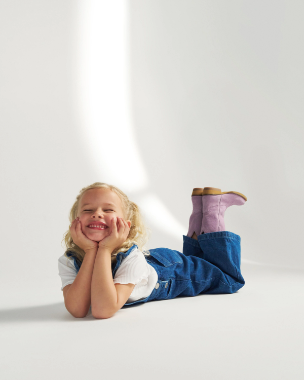 A young child with blonde hair, wearing a white shirt, blue overalls, and pink boots, lies on the floor smiling with hands under their chin.