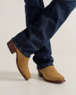 A person wearing dark blue jeans and tan suede cowboy boots stands with their feet crossed on a light background.