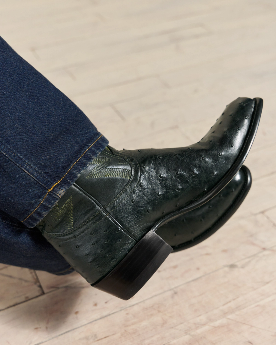 A close-up of someone wearing dark green cowboy boots made from textured leather and dark blue jeans on a light wooden floor.