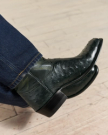 A close-up of someone wearing dark green cowboy boots made from textured leather and dark blue jeans on a light wooden floor.