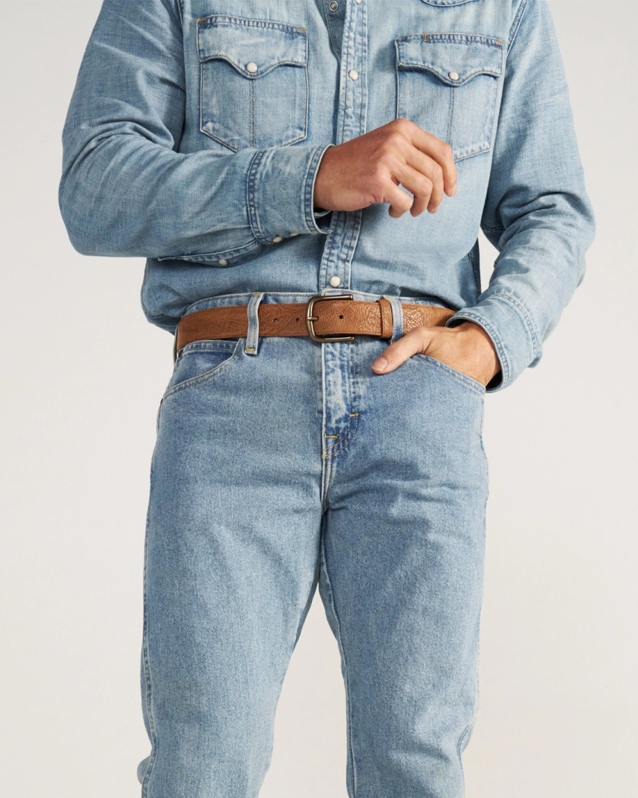 A person wearing a light blue denim shirt tucked into matching light blue jeans, secured with a brown leather belt.