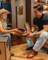 A woman in a cowboy hat speaks to a seated man holding a cowboy boot in a store, with boots and boxes around them.
