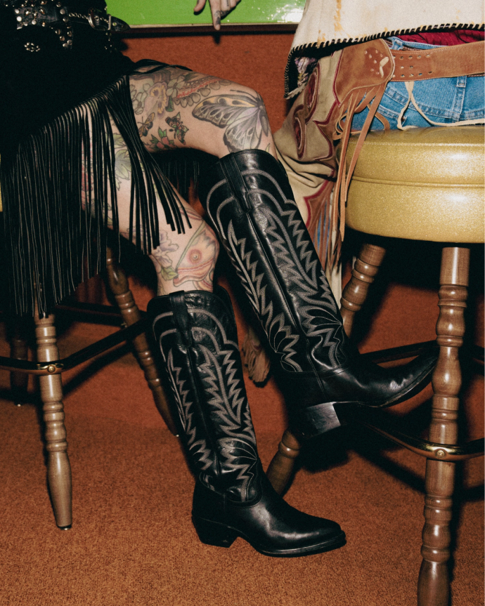 Two people sitting on stools wearing fringed clothing and tall black cowboy boots, with one person's tattooed legs visible.