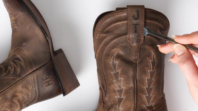 A hand embosses the initials "JT" onto a brown leather cowboy boot; another boot with the initials "HF" is visible nearby.
