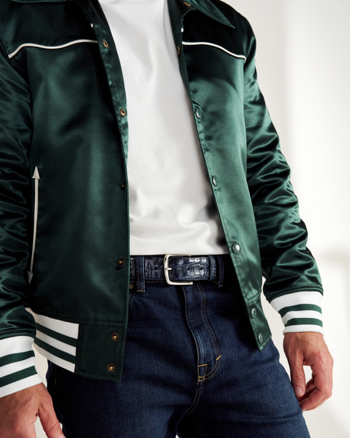 Person wearing a dark green satin jacket with white accents over a white shirt, paired with blue jeans and a black belt.