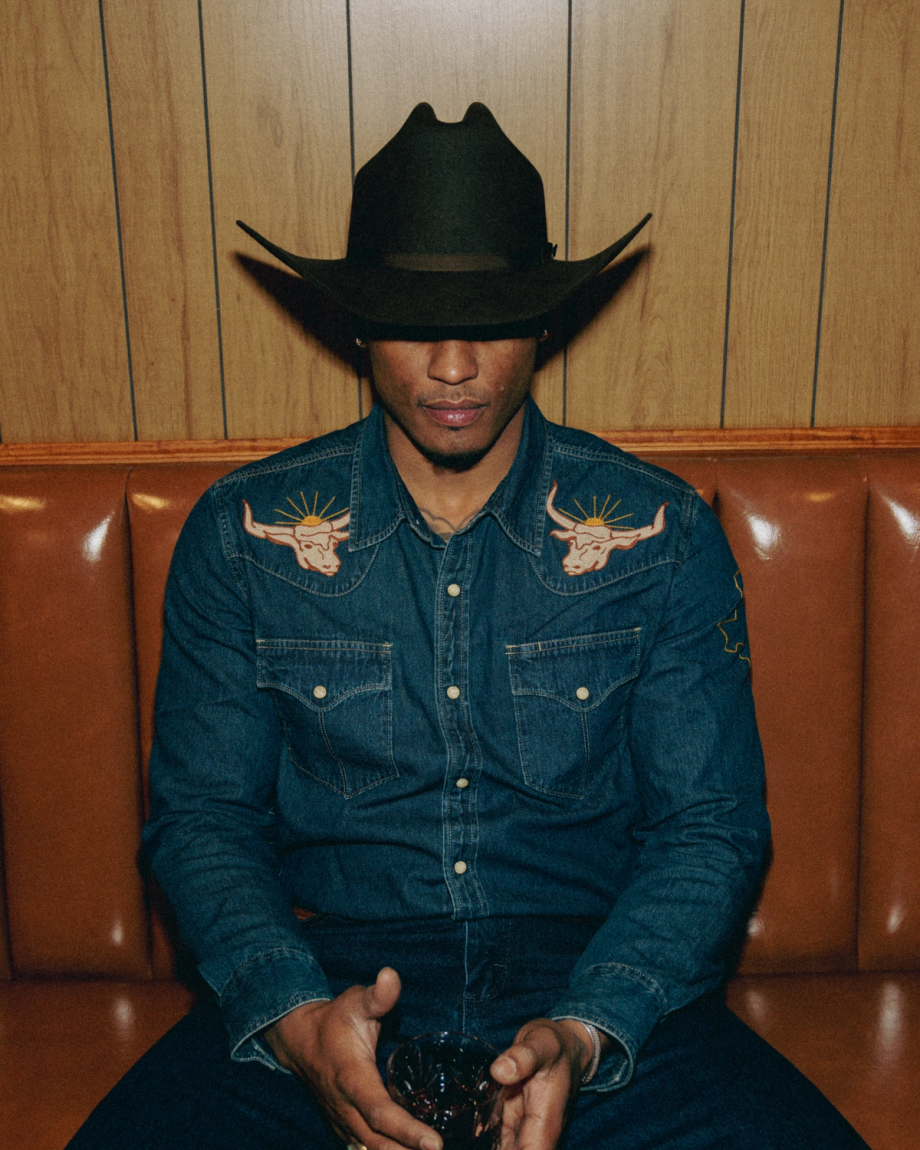 A person sits on a brown leather bench in a denim shirt with embroidered bull designs, wearing a dark cowboy hat and holding an item in their hands. Wood-paneled wall in the background.