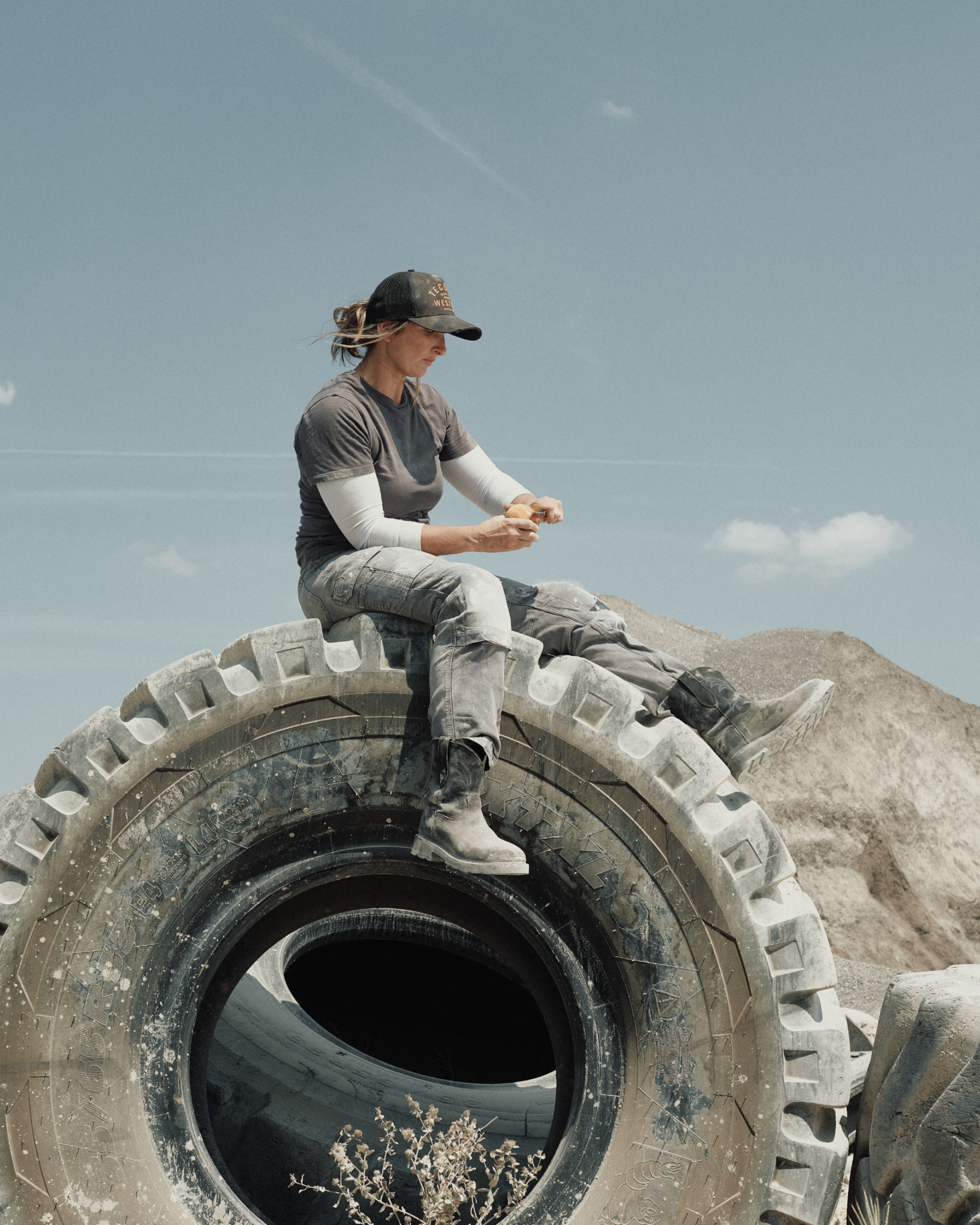 Person wearing work clothes and a hat sits on a large tire, eating a snack, with dirt mounds and a blue sky in the background.
