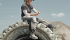 Person wearing work clothes and a hat sits on a large tire, eating a snack, with dirt mounds and a blue sky in the background.