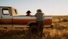 Two people in cowboy hats talk beside a vintage brown and white pickup truck parked in a dry grassland at golden hour.