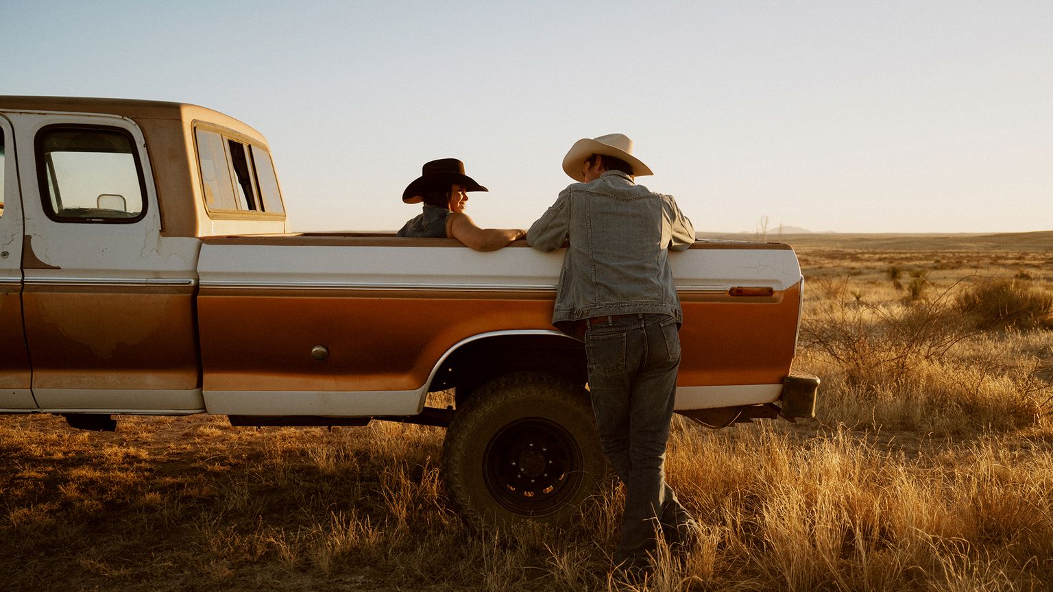 Two people in cowboy hats talk beside a vintage brown and white pickup truck parked in a dry grassland at golden hour.