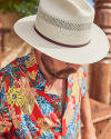 Man wearing a beige hat and a colorful floral shirt looks down while standing outdoors.