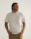 A man with braided hair and tattoos wears a light gray t-shirt and khaki pants, standing against a plain background with his hands in his pockets, looking to the side and smiling.
