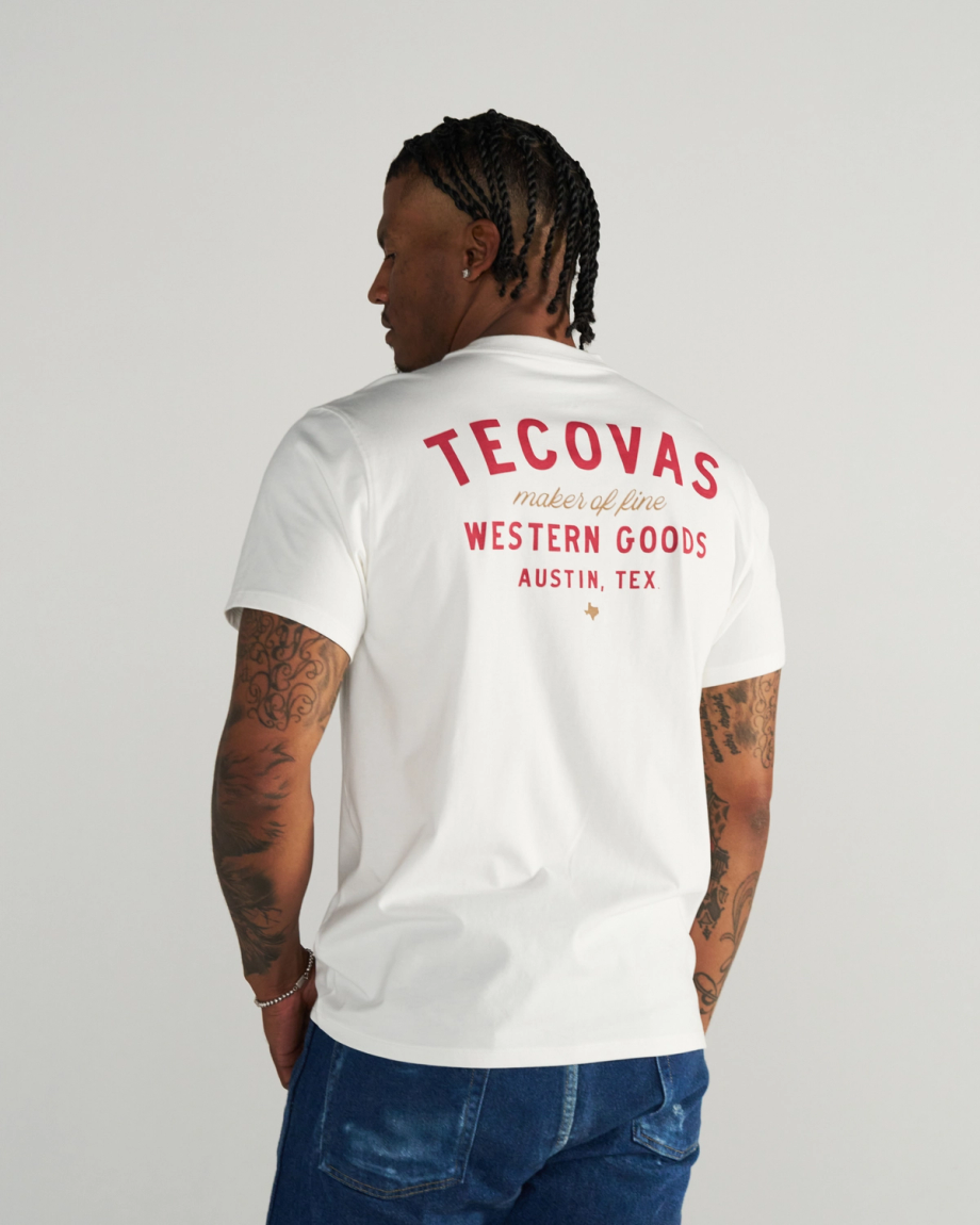 A person with braided hair wears a white T-shirt with red text reading “Tecovas maker of fine Western Goods Austin, Tex” on the back, standing against a plain background.