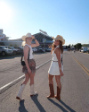 Two women wearing cowboy hats and boots stand on an empty road by a parking lot, turning to face the camera, with a stadium visible in the background.