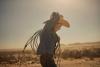 A child in a cowboy hat and denim shirt holds a coiled rope against a hazy desert backdrop.
