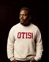 Man in a cream sweatshirt with "OTIS!" written in red, stands against a dark background, looking to the side.