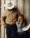 An adult and a child in cowboy hats and western attire stand against a corrugated metal wall; the child holds a hat and touches their forehead.