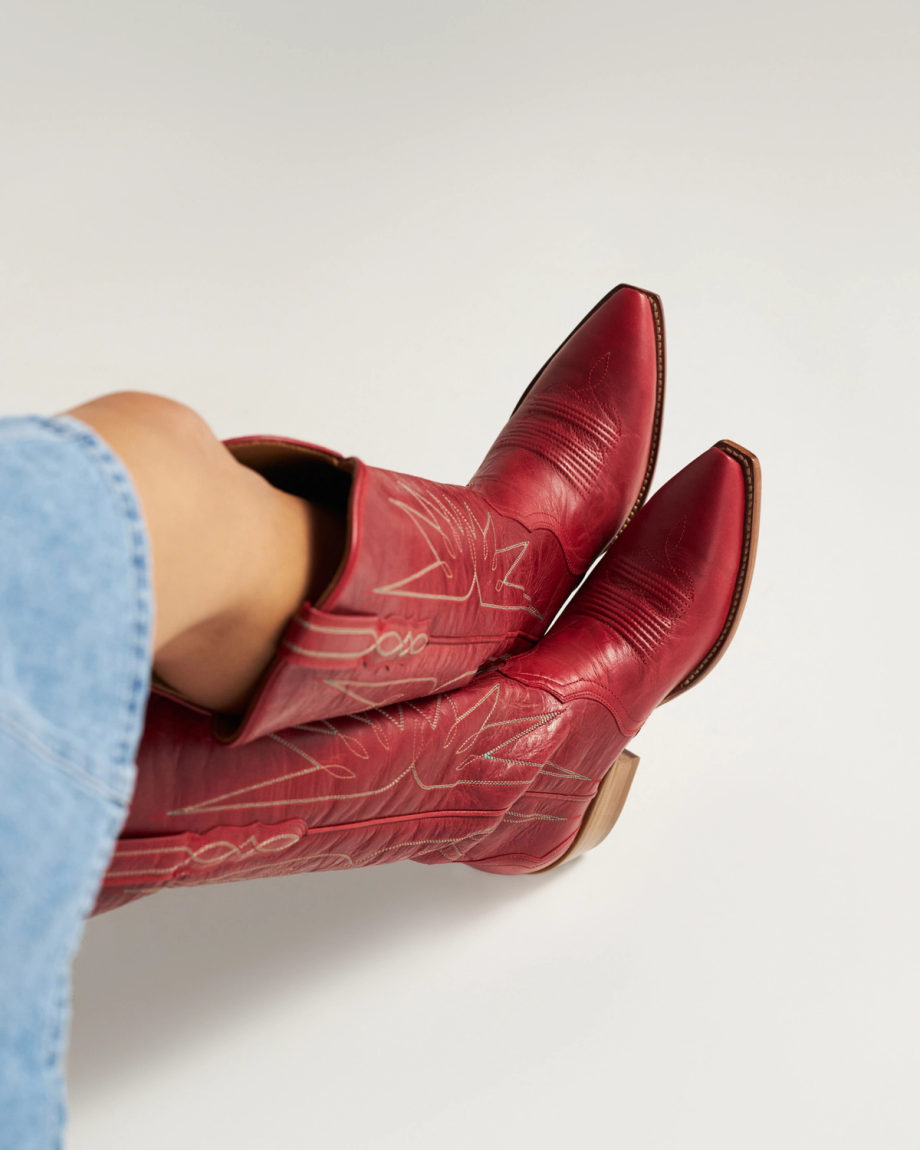 The Loretta in Ruby Goat. Person wearing red cowboy boots with decorative stitching, partially covered by a light blue denim garment, stands confidently like a Ruby Goat against a plain light background.