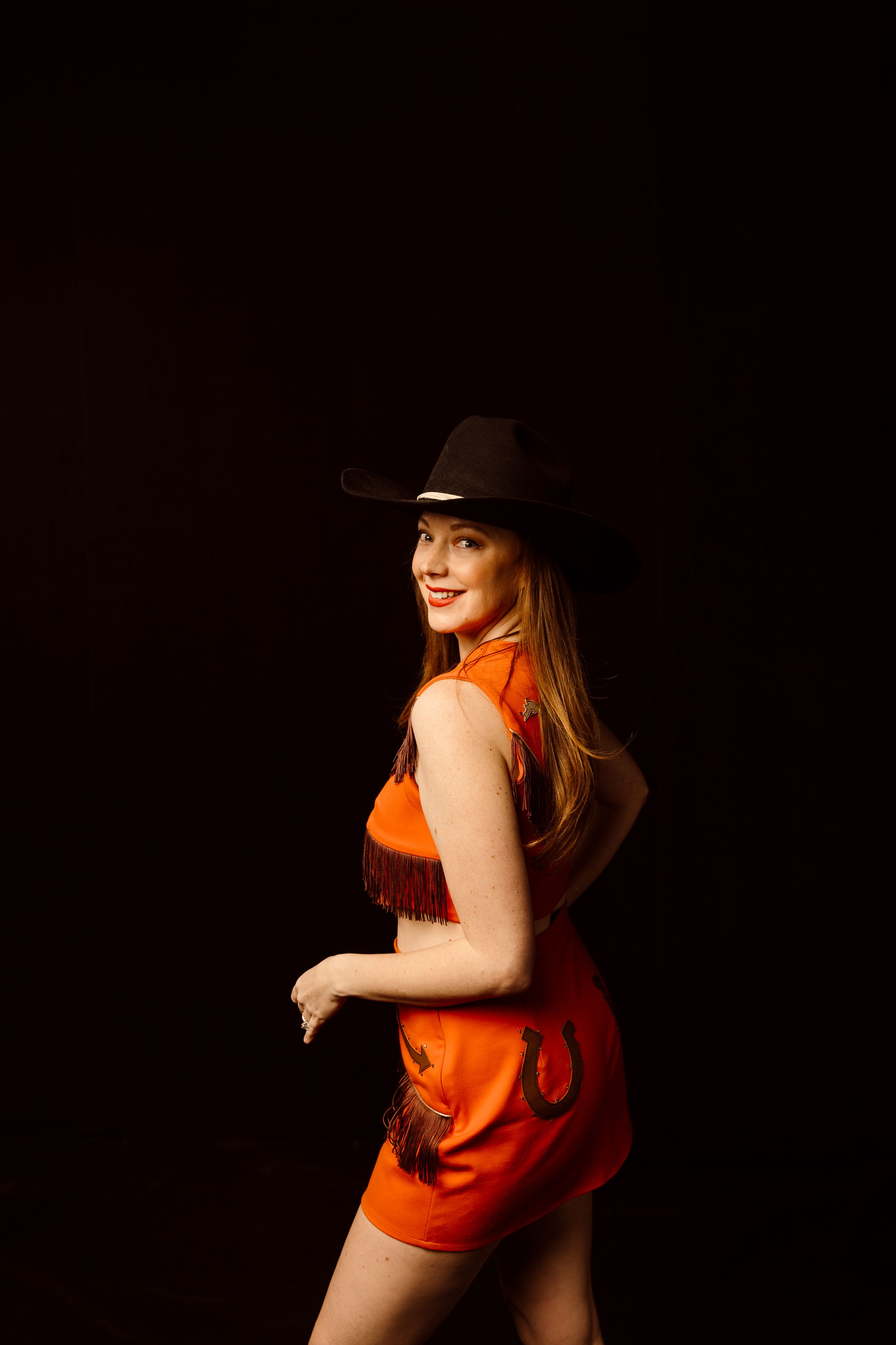 Person in a red fringed outfit and black cowboy hat, smiling against a dark background.