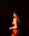 Person in a red fringed outfit and black cowboy hat, smiling against a dark background.
