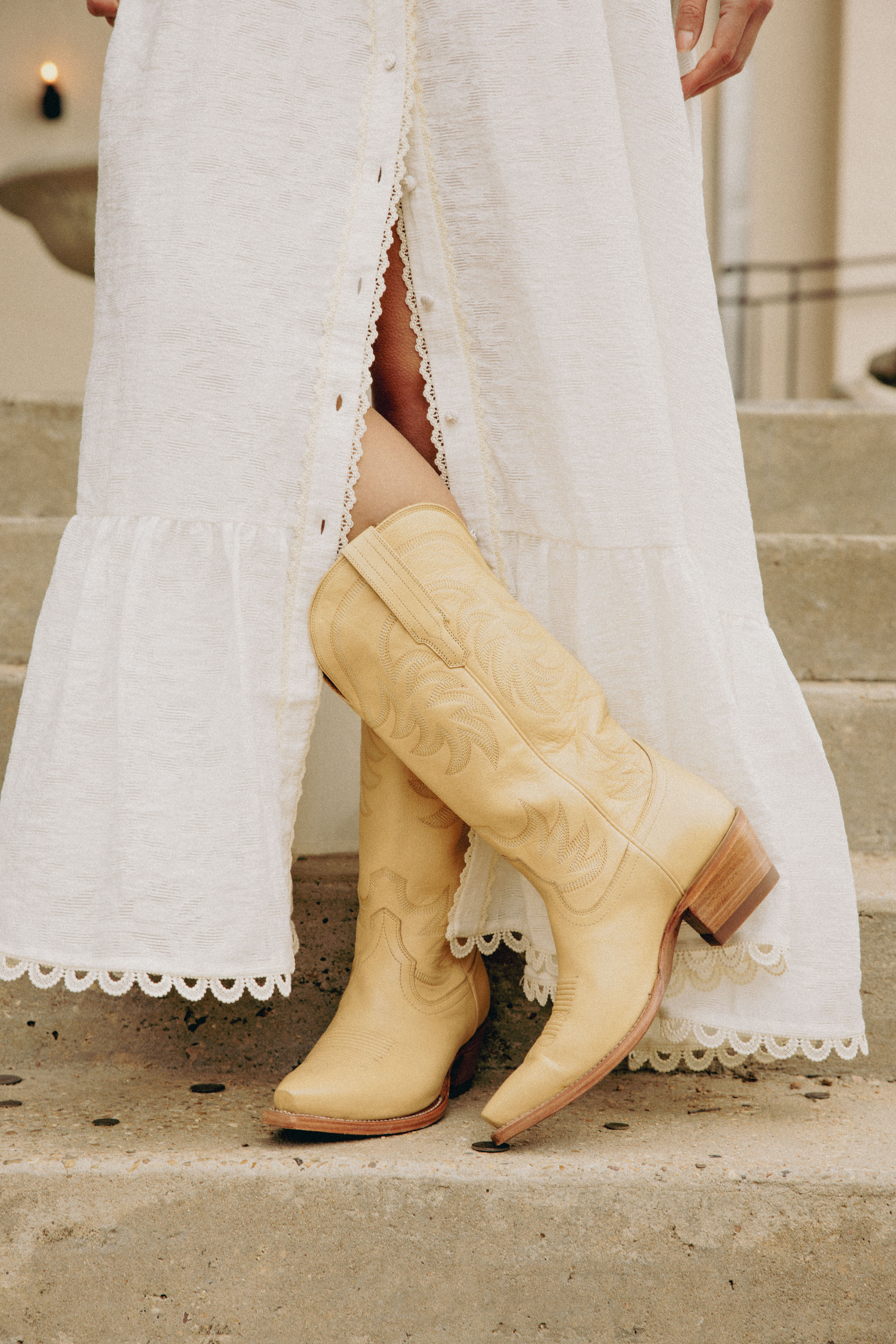 Person in a long white dress stands on steps, wearing light tan cowboy boots with embroidery detailing.