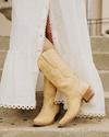 Person in a long white dress stands on steps, wearing light tan cowboy boots with embroidery detailing.