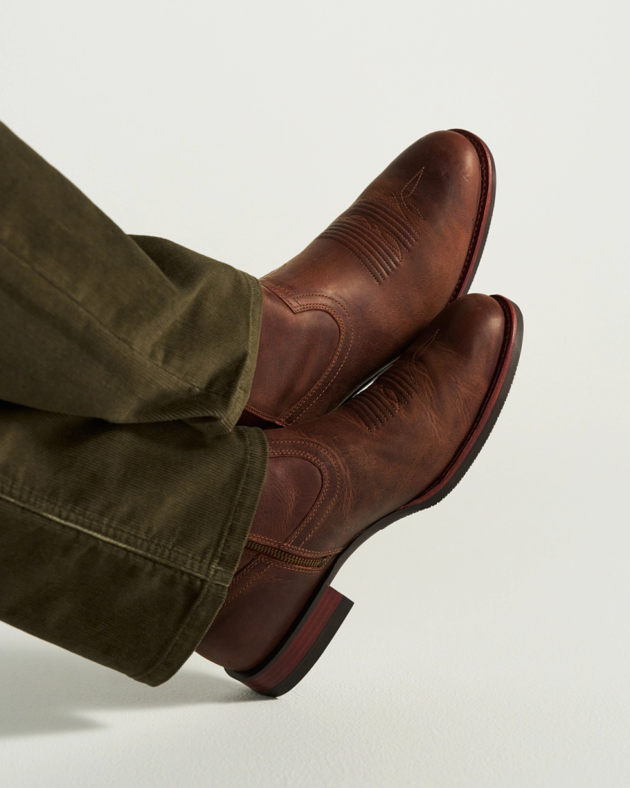 The Wayne in Maplewood Cowhide. Person wearing olive green pants and brown leather cowboy boots, featuring rich Maplewood Cowhide, shown from the side with feet crossed and resting on a light surface.