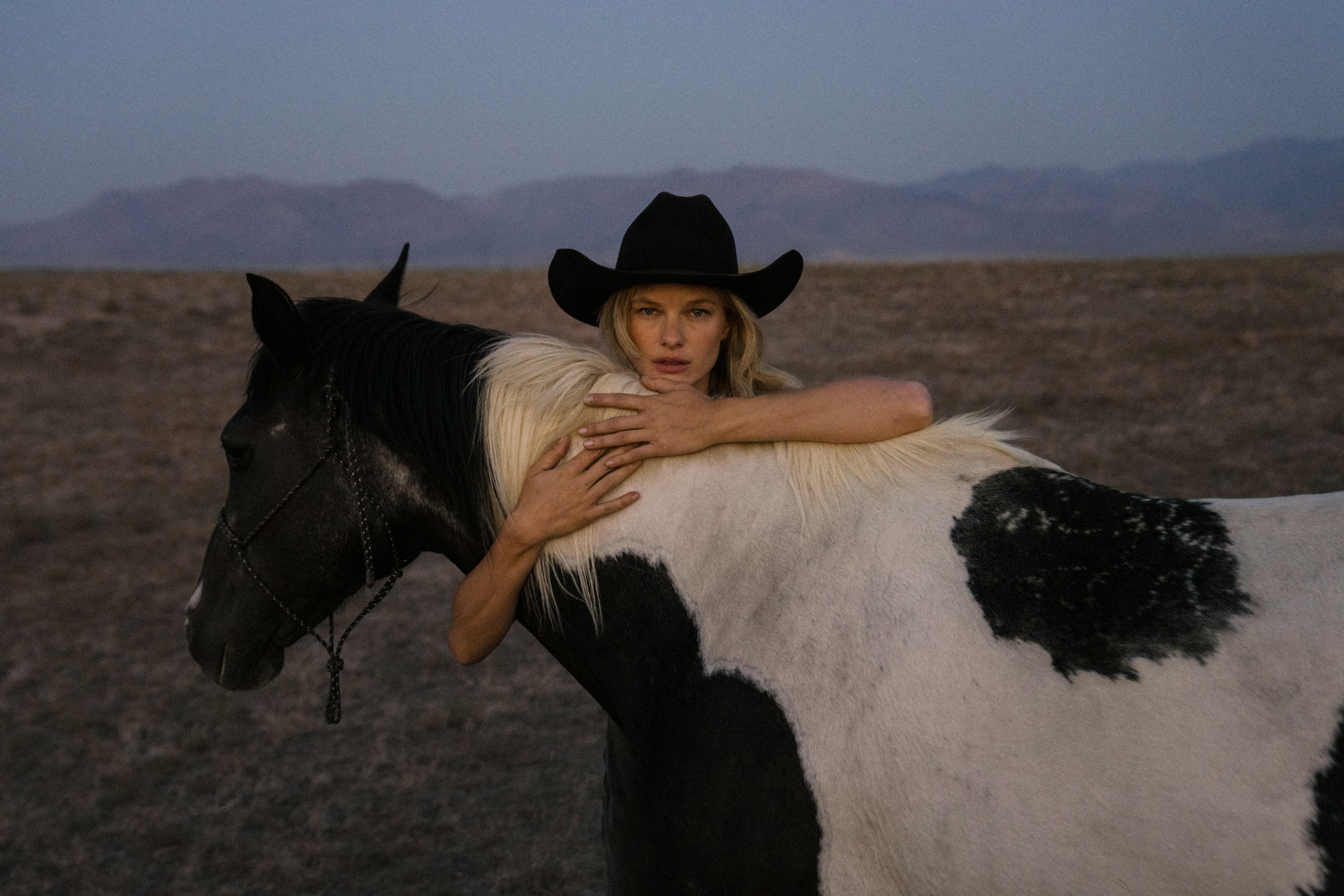 A person in a black cowboy hat embracing a black and white horse in a desert landscape.