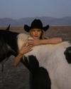 A person in a black cowboy hat embracing a black and white horse in a desert landscape.
