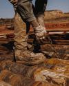 A worker wearing gloves and muddy boots secures chains around large metal pipes outdoors at a construction or industrial site.